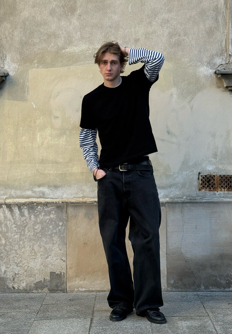 Jeune homme portant une chemise à manches longues rayée sous un T-shirt noir, un pantalon large noir et des chaussures en cuir, debout contre un mur beige texturé.