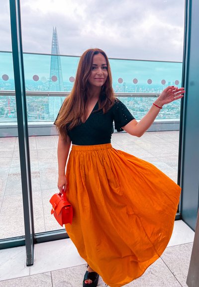 Mujer con cabello largo castaño, vestida con una blusa negra y una falda naranja, se encuentra dentro de una plataforma de observación de vidrio con vista al horizonte de la ciudad.
