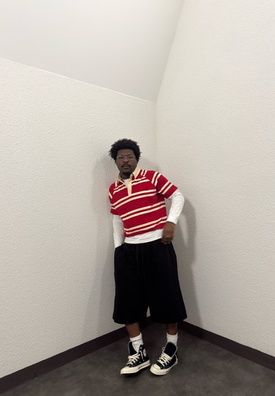 Hombre con gafas que lleva una camisa roja de rayas sobre una camisa blanca de manga larga, pantalones cortos negros y zapatillas negras de caña alta, se apoya contra una pared blanca de esquina.