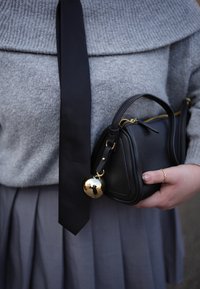 Μαύρη δερμάτινη τσάντα με δομημένο σχήμα, με χρυσή μπάλα ως λεπτομέρεια, κρατιέται μπροστά σε ένα γκρίζο πουλόβερ. Μερικώς ορατή γκρίζα πλισέ φούστα.