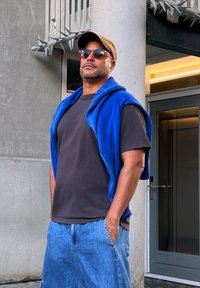 Gilet blu lavorato a maglia su una maglietta nera, pantaloni corti di jeans e occhiali da sole. Lo sfondo presenta un muro di cemento e una porta con vetro.