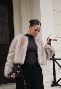 Κοντό, ανοιχτό καφέ ψεύτικο γούνινο μπουφάν με φαρδιά μανίκια, φορεμένο πάνω από ένα σκούρο λαιμόκοψη. Το μοντέλο κρατάει γυαλιά ηλίου και μια σκούρα τσάντα χιαστί.