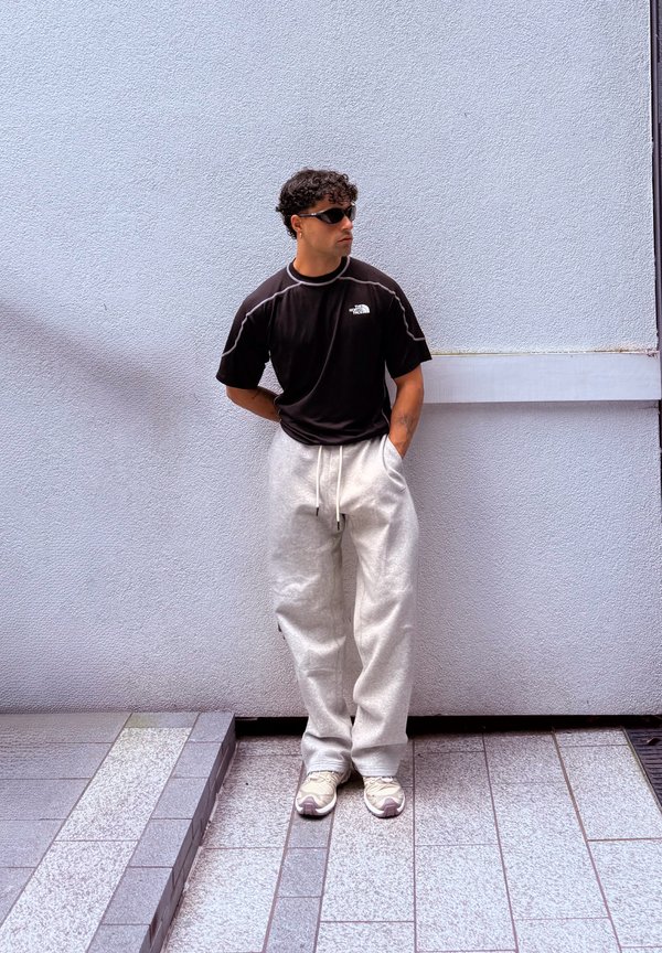 Hombre con cabello rizado y gafas de sol se apoya contra una pared blanca texturizada, viste una camiseta negra, pantalones deportivos gris claro y zapatillas beige con las manos en los bolsillos.