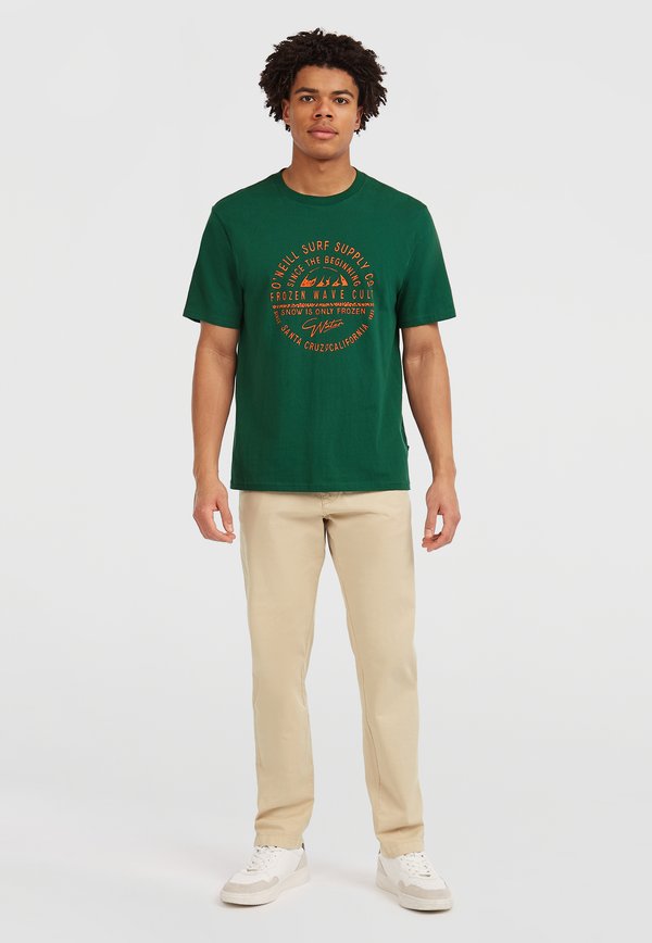 T-shirt en coton vert avec un texte graphique orange et des images de montagnes, associé à un pantalon beige et des baskets blanches.
