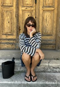 Mulher com uma camisa de mangas longas listrada, óculos de sol e chinelos sentada em escadas de pedra em frente a portas de madeira desgastadas, ao lado de uma bolsa preta.