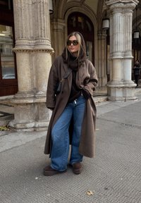 Donna con un cappotto marrone oversize, maglione scuro, jeans a gamba larga, occhiali da sole e scarpe marroni che stands vicino a colonne di pietra su una strada cittadina.