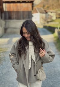 Béžový vlnený kabát s nadrozmerným strihom, doplnený textúrovaným krémovým šálom. Malá vzorovaná kabelka a svetlé nohavice dopĺňajú outfit.