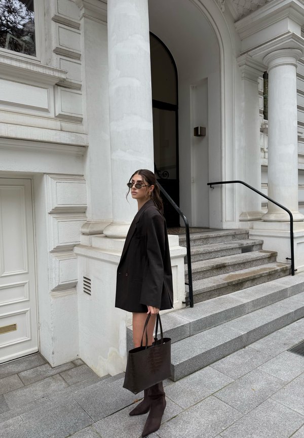 Musta ylisuurena bleiseri, polvimittainen hame, tumma kuviollinen käsilaukku, polven korkeudelle ulottuvat kärkitossut, seisomassa valkoisten pylväiden ja kivisten portaiden vierellä.