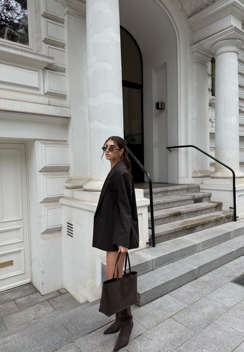Blazer negro de gran tamaño, falda a la altura de la rodilla, bolso oscuro estampado, botas de tacón alto hasta la rodilla, de pie junto a columnas blancas y escaleras de piedra.