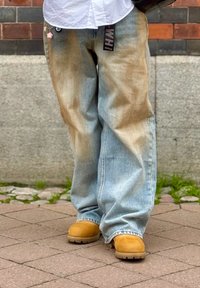 Lyseblå denim jeans med en afslappet pasform, der viser slidte områder, parret med brune støvler. Et sort mærke hænger fra jeansene.
