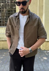 Man met zwarte zonnebril, een bruin overhemd over een wit T-shirt, zwarte broek en zilveren horloge, staande op een geplaveide loopbrug.