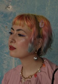 Cabello corto con un degradado de rosa, amarillo y azul; lleva una blusa rosa; acentuado por joyas coloridas y un clip de pelo rosa.