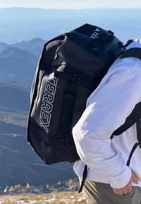 Personne portant un sweat-shirt blanc et un pantalon vert, portant un grand sac à dos noir TERREX sur une montagne avec des sommets en couches en arrière-plan.