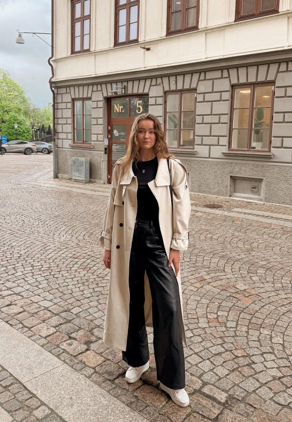 Kvinde står på en brostensbelagt gade iført en lang beige trenchcoat, sort top, sorte bukser og hvide sneakers med et neutralt udtryk.