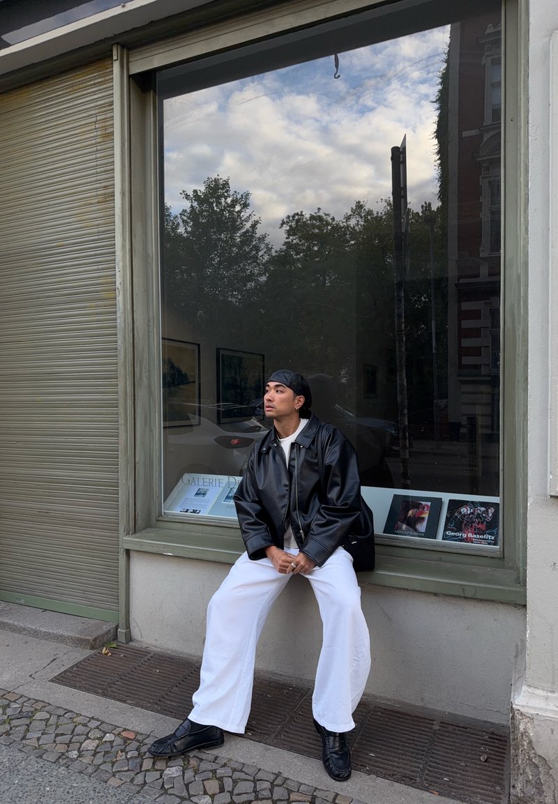 Personne portant une veste en cuir noire et un pantalon blanc, assise sur un rebord sous une grande fenêtre reflétant des arbres et un ciel nuageux.