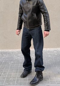 Άτομο που φοράει μαύρο δερμάτινο μπουφάν, σκούρο μπλε τζιν και μαύρες δερμάτινες μπότες, στέκεται σε πλακόστρωτο πεζοδρόμιο μπροστά σε μπεζ τοίχο.