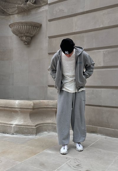 Modelo vistiendo una sudadera gris oversize, camiseta gris claro y pantalones de chándal grises holgados, complementados con zapatillas blancas y una gorra negra.