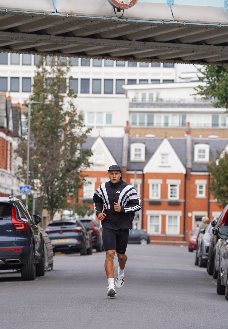 Giovane uomo che corre su una strada della città sotto un ponte, indossando un cappello nero, una giacca bianca e nera, pantaloncini neri e sneakers bianche.