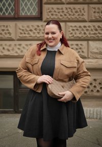 Femme souriante aux cheveux roux, portant une veste beige et une robe noire, tenant un sac à main de créateur marron contre un mur en pierre texturé.