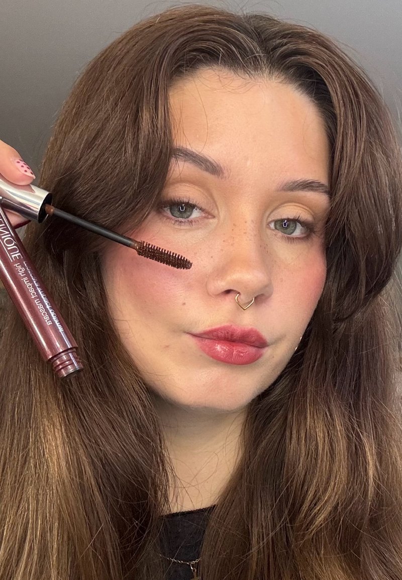 Tubetto di mascara con pennello, confezione bordeaux, tappo argento. Modella con capelli lunghi e castani, pelle liscia, trucco delicato e anello al setto nasale.