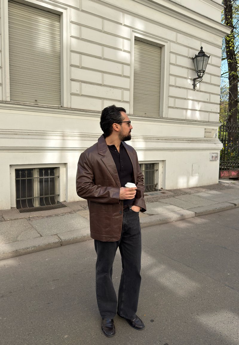 Homme en veste en cuir marron et pantalon noir tenant une tasse de café, debout dans une rue de la ville près d'un bâtiment blanc aux fenêtres fermées.