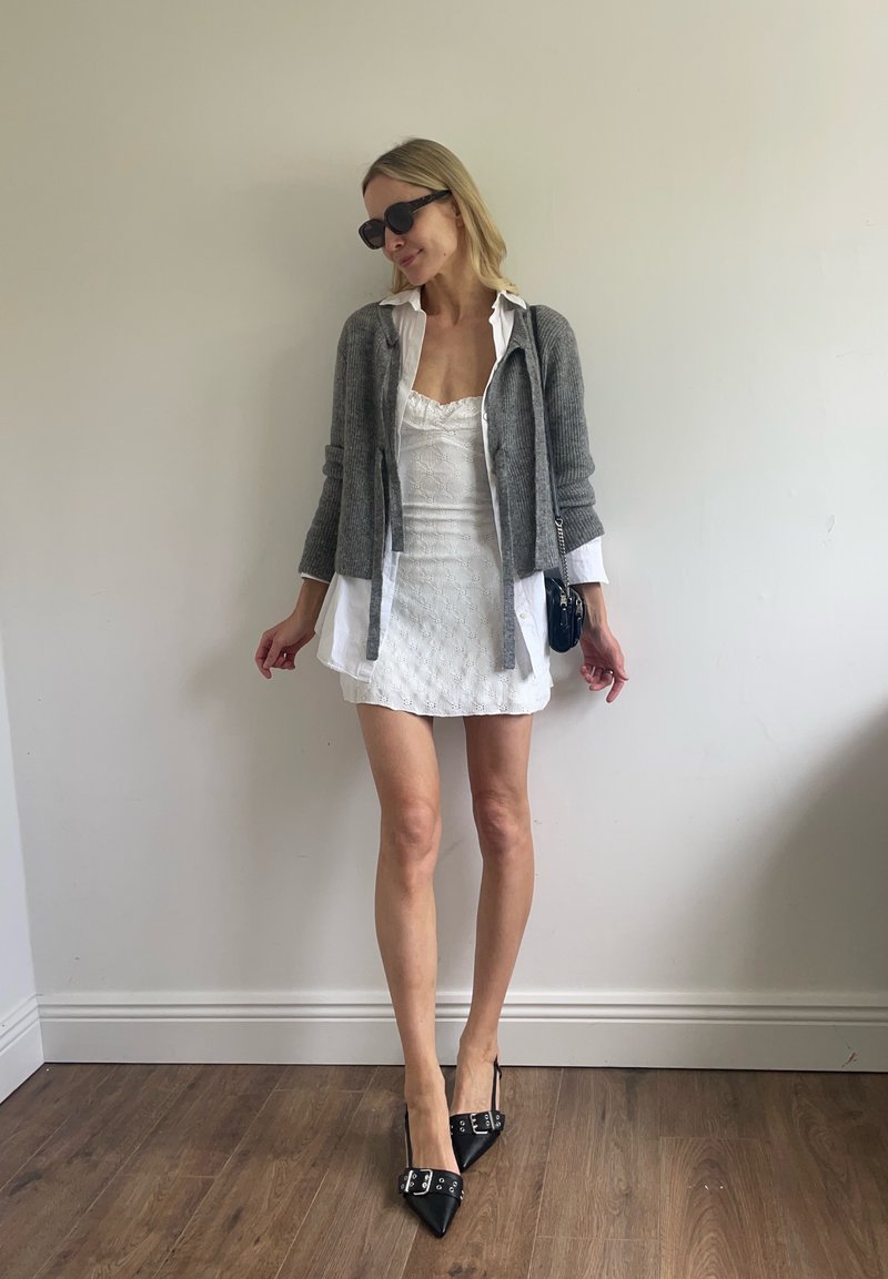 Femme portant une mini-robe blanche avec une chemise blanche ouverte et un cardigan gris, lunettes de soleil noires, talons pointus noirs et un petit sac à épaule noir.
