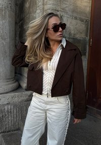 Kvinde med langt blond hår iført store mørke solbriller, en mørk kort jakke, hvid stribet skjorte og hvide bukser, stående ved stensøjler.