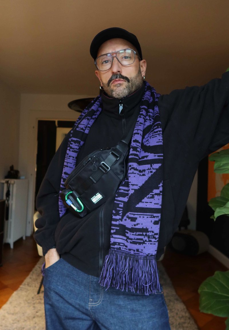 Homme portant une casquette noire, des lunettes, une veste noire, une écharpe à motifs violets et un sac bandoulière, debout à l'intérieur avec une main dans la poche.
