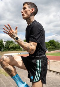 Calças curtas de atletismo pretas com riscas teal, uma camisola preta ajustada e meias azuis com padrão. Tatuagens cobrem os braços e um relógio inteligente está preso ao pulso.