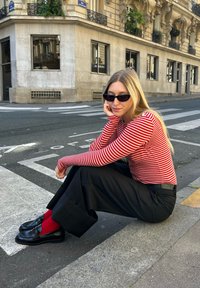 Junge Frau mit blonden Haaren, schwarzen Sonnenbrillen, einem rot-weißen gestreiften Oberteil, schwarzen weiten Hosen und schwarzen Loafers, die auf dem Bordstein einer Stadtstraße sitzt.