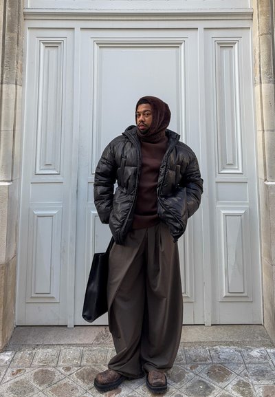 Chaqueta acolchada negra, suéter marrón, pantalones marrones holgados, zapatos oscuros y un bolso tote negro, de pie frente a una puerta blanca con paneles.