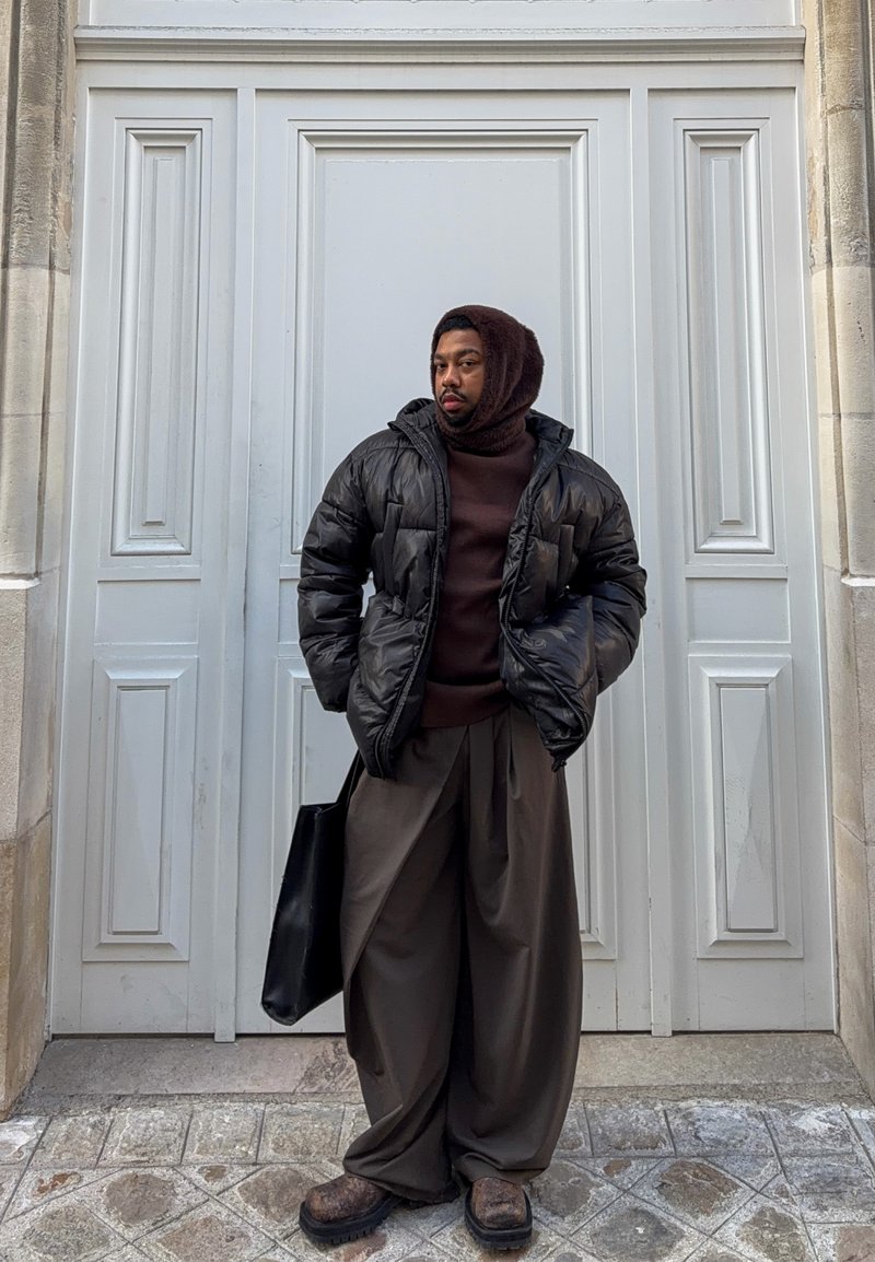 Schwarze Steppjacke, brauner Pullover, lockere braune Hose, dunkle Schuhe und eine schwarze Tragetasche, stehend vor einer weißen Tür mit Paneelen.