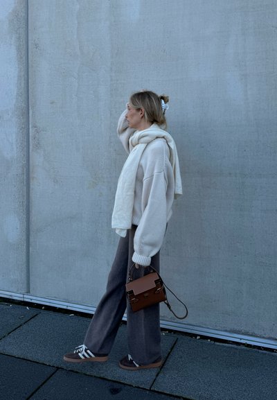 Mujer con un acogedor suéter blanco y bufanda, pantalones anchos grises, zapatillas marrones y bolso marrón caminando junto a una pared de concreto.