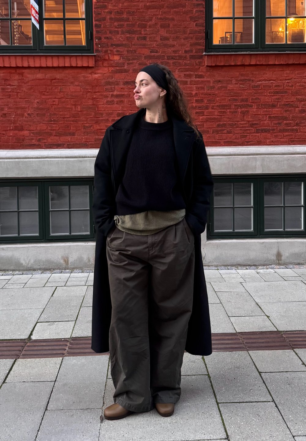 Fekete hosszú kabát fekete bordázott pulóver fölött, olívazöld, bő szárú nadrág texturált derékrésszel, és barna cipők. Piros téglafal a háttérben.
