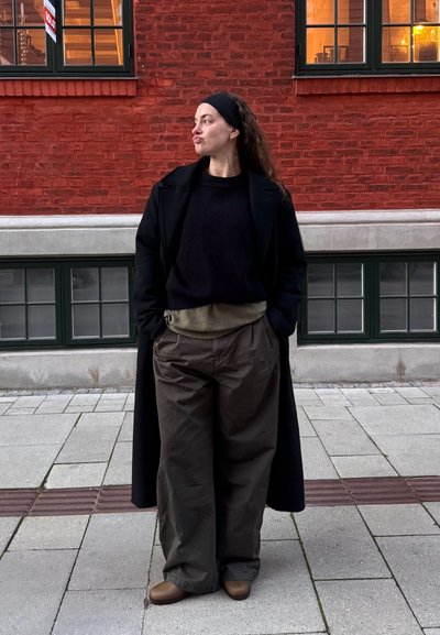 Abrigo largo negro sobre un suéter negro de canalé, pantalones anchos verde oliva con una pretina texturizada y zapatos marrones. Fondo de pared de ladrillo rojo.