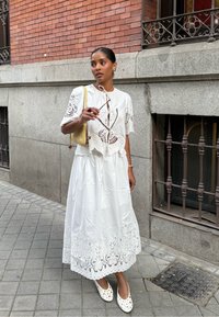 Donna in abito di pizzo bianco e ballerine che tiene occhiali da sole e una borsa gialla a spalla, in piedi su un marciapiede piastrellato vicino a un edificio in mattoni e pietra.