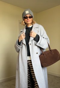 Grey wool coat with a notched collar, black knitted jumper, brown striped trousers, grey beanie, and brown handbag. Accessories include several rings.