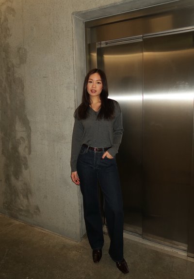 Mujer con suéter gris y jeans oscuros, con la mano en el bolsillo, de pie junto a una puerta de ascensor metálica cerrada en un pasillo de hormigón.