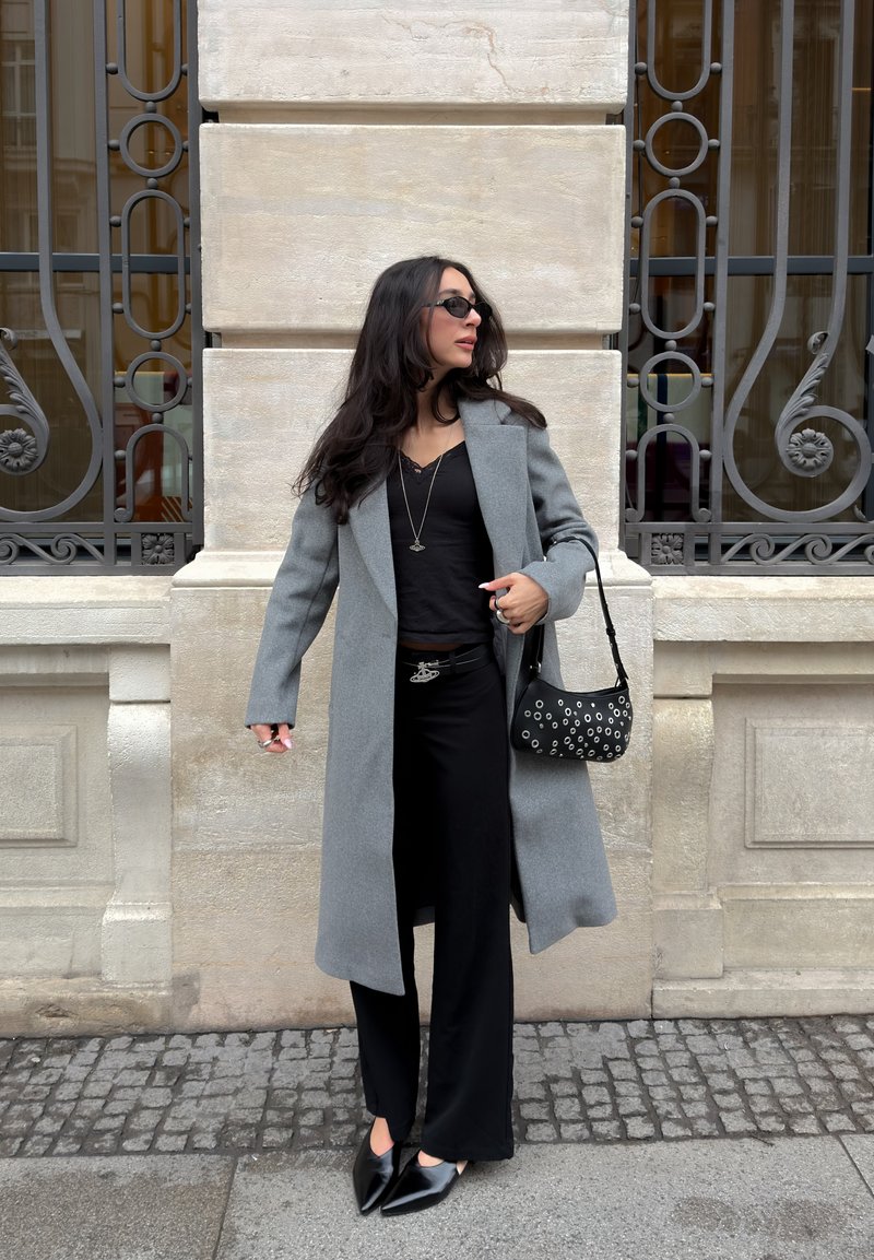 Femme en manteau gris, tenue noire et lunettes de soleil, debout sur le trottoir de la ville devant un bâtiment en pierre avec des grilles de fenêtres décoratives en fer.