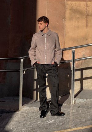 Joven de pie al aire libre bajo la luz del sol, llevando una chaqueta beige con cremallera, pantalones negros con rayas finas y zapatos negros, con las manos en los bolsillos.