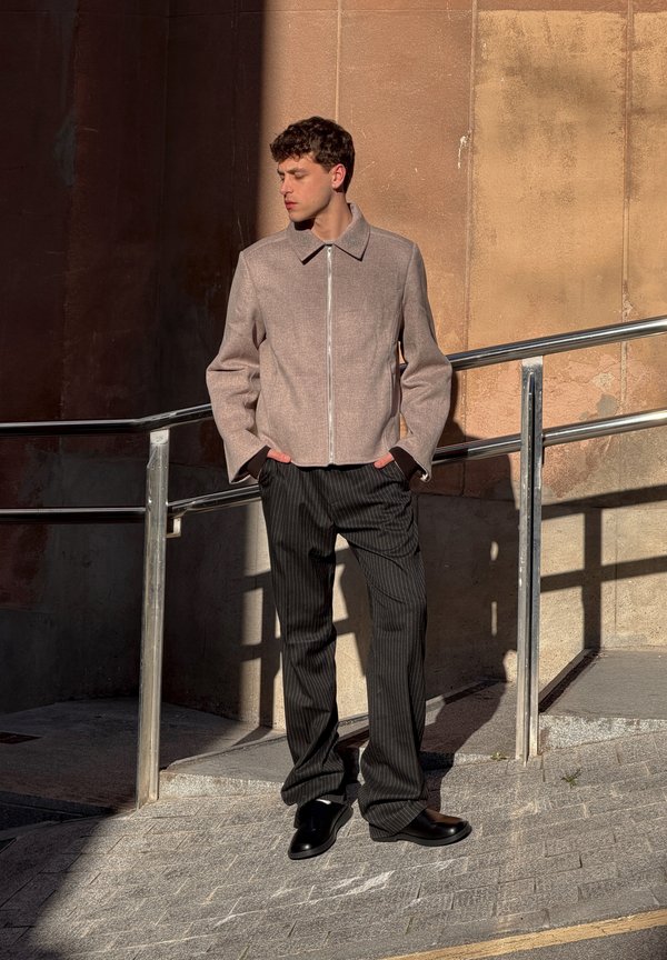 Νεαρός άνδρας στέκεται έξω στον ήλιο, φορώντας μπεζ ζακέτα με φερμουάρ, μαύρο παντελόνι με ρίγες και μαύρα παπούτσια, με τα χέρια στις τσέπες.