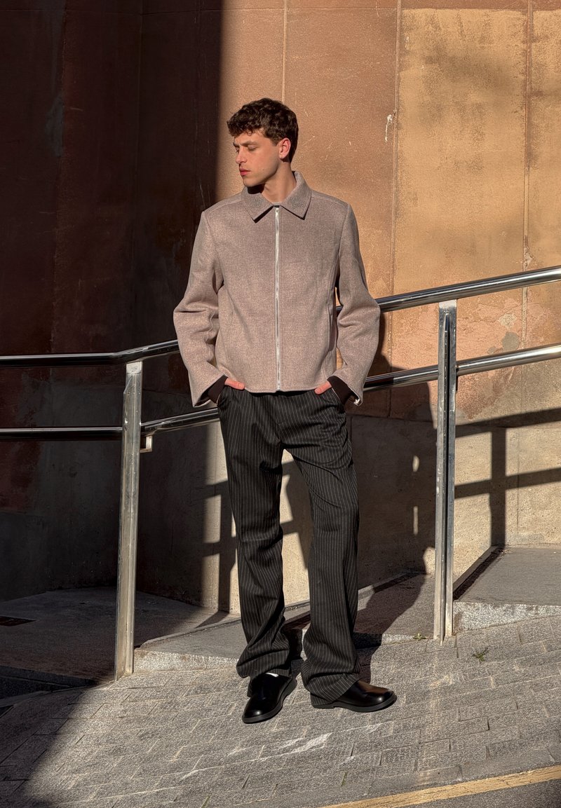 Jeune homme debout en plein air sous la lumière du soleil, portant une veste beige zippée, un pantalon noir à fines rayures et des chaussures noires, les mains dans les poches.