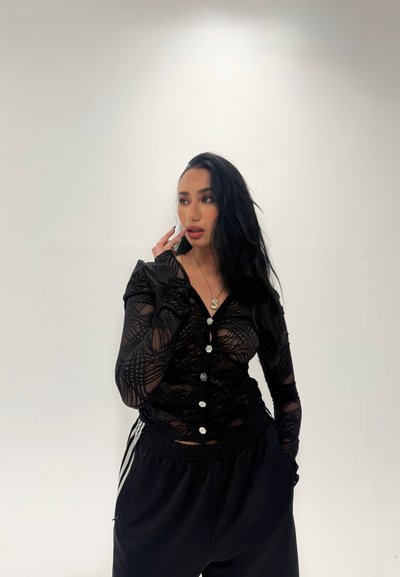 Top de malla negra de manga larga con patrones intrincados y cierre de botones, combinado con pantalones deportivos negros con rayas blancas a los lados.