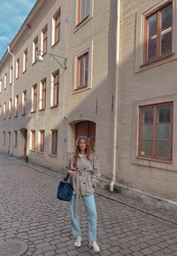 Svetlo béžový trenčkot s viazaným pásom, svetlomodré rovné džínsy a biele tenisky. Tmavá taška je prehodená cez jedno rameno.