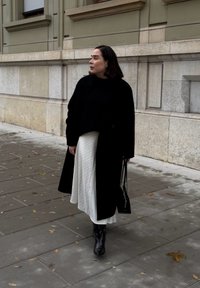 Sort uldfrakke over en sort top og hvid, stribet midinederdel. Sorte støvler med hæl og en lille sort håndtaske. Fortov med faldne blade.