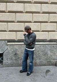 Man i grå tröja, blå jeans och svarta skor som justerar en brun keps, stående på trottoar mot en texturerad beige och grå stenvägg.