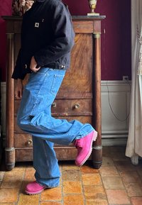 Svart quiltet jakke med frontlomme, blå vide jeans og lys rosa ankelstøvletter med teksturerte såler, posisjonert mot en trebakgrunn.