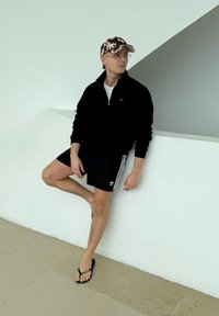 Νέος άνδρας που φοράει μαύρο πουλόβερ Nike, σορτς Adidas, σαγιονάρες και καπέλο με σχέδιο αγελάδας, ακουμπά σε λευκό τοίχο σε εσωτερικό χώρο.