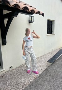 White t-shirt with red text, patterned grey snake-print trousers, pink platform sandals, and a small white handbag, standing against a textured wall.