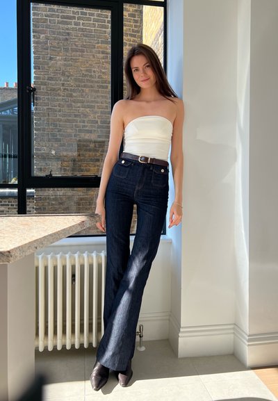 Jeans de mezclilla oscuros de pierna acampanada con cintura alta, combinados con una blusa sin mangas blanca. Incluye un cinturón marrón, botas negras pulidas hasta el tobillo y un fondo neutro.